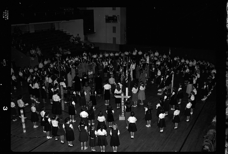 Group of unidentified people gathered in a room for the Blue Bird Fly-Up. Men, women, and mostly children are all standing in a circle. Decorated poles and flags are visible.