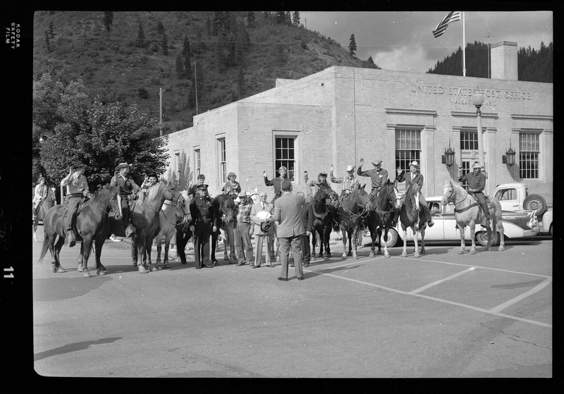 Group of people lined up with many on horseback and some raising their hands. They are standing in the parking lot of the Wallace, Idaho post office with the building, cars, and trees visible in the background.