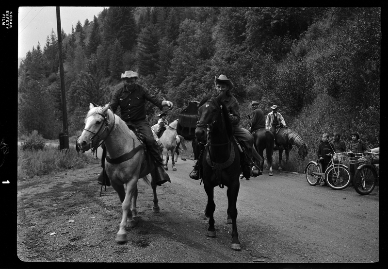 Two horse back riders from Pony Express are the focus, with other horse riders in the background with some children on bikes. They are riding on a dirt road and one of the riders is reaching out for the other.