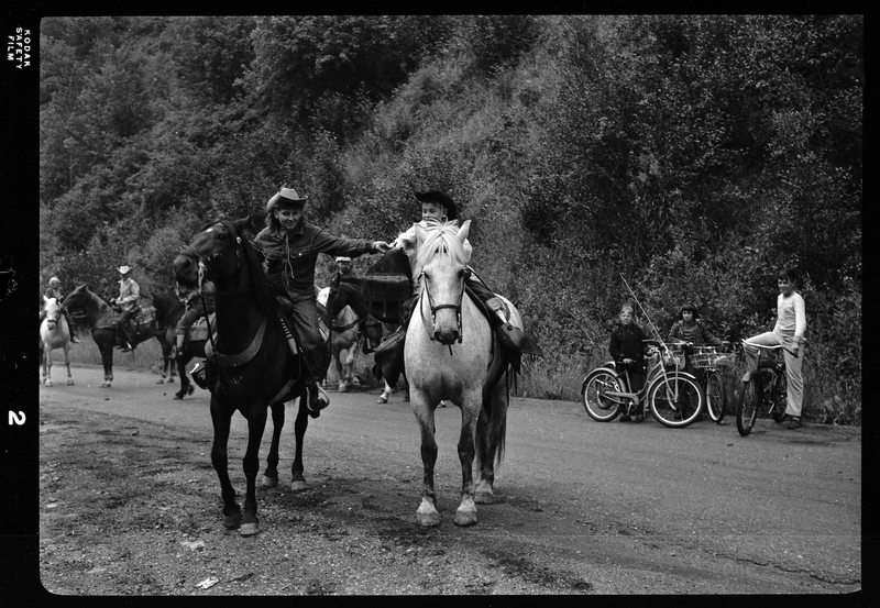 Two horse back riders from Pony Express are the focus, with other horse riders in the background with some children on bikes. They are riding on a dirt road and the riders are reaching out for each other.