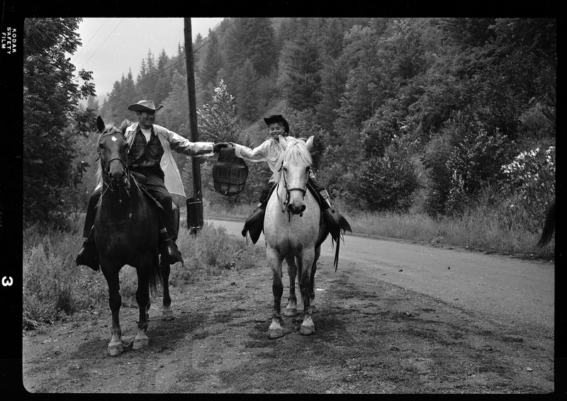 Two horse back riders, a man and a child, from Pony Express on a dirt road. They are both holding up the same bag between them. 
