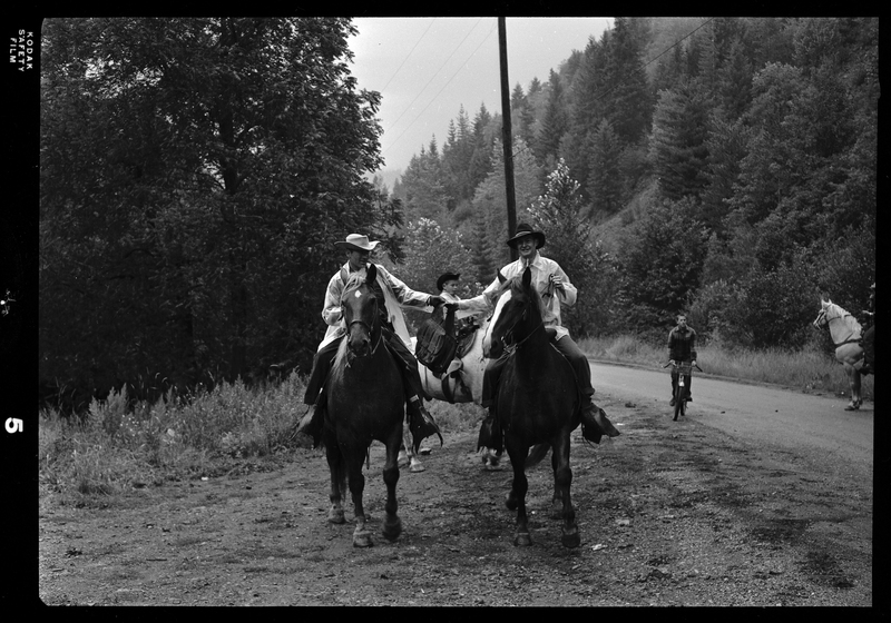 Two horse back riders, both men, from Pony Express on a dirt road with other horse back riders visible in the background as well as a child on a bicycle. They are both holding up the same bag between them. 