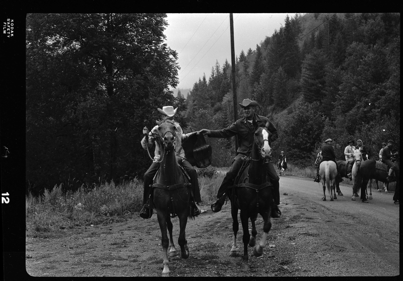 Two horse back riders, both men, from Pony Express on a dirt road with other horse back riders visible in the background. They are both holding up the same bag between them. 