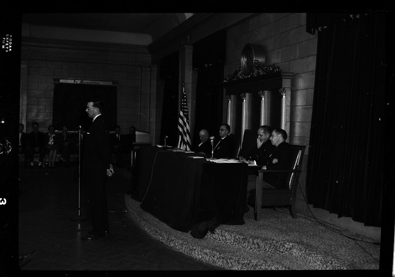 Panel of men sitting behind long desk with one man standing in front of them. The man standing is in front of a microphone and seemingly addressing the crowd. There is an American flag visible in the background.