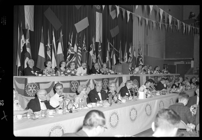 Rotary International Club Convention panel that includes Gordon Beaton, Greek Wells, Guy M'Loughlin, Bill Featherstone, and Bob Green. Various men and women are seated in two rows with dinner ware on the tables in front of them. There are banners hanging from the ceiling and various flags along the back wall.