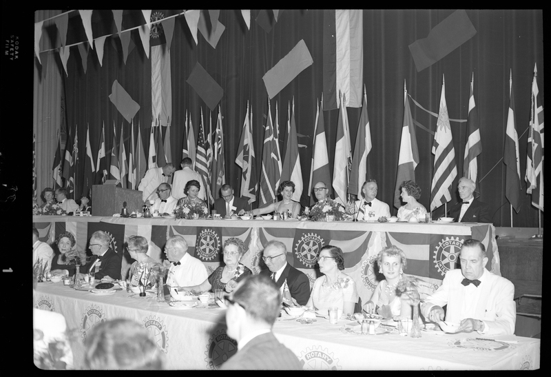 Rotary International Club Convention panel that includes Gordon Beaton, Greek Wells, Guy M'Loughlin, Bill Featherstone, and Bob Green. Various men and women are seated in two rows with dinner ware on the tables in front of them. There are banners hanging from the ceiling and various flags along the back wall.
