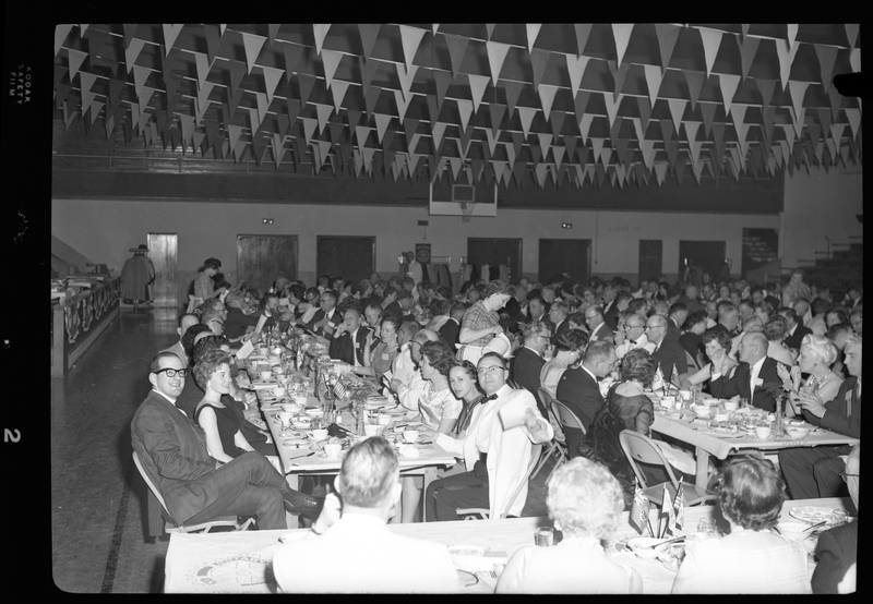 Crowd of unidentified men and women gathered in a room while eating for the Rotary International Club Convention. Some people are looking at the camera while most are interacting with each other. Banners have been hung across the ceiling and there is a basketball hoop in the background.
