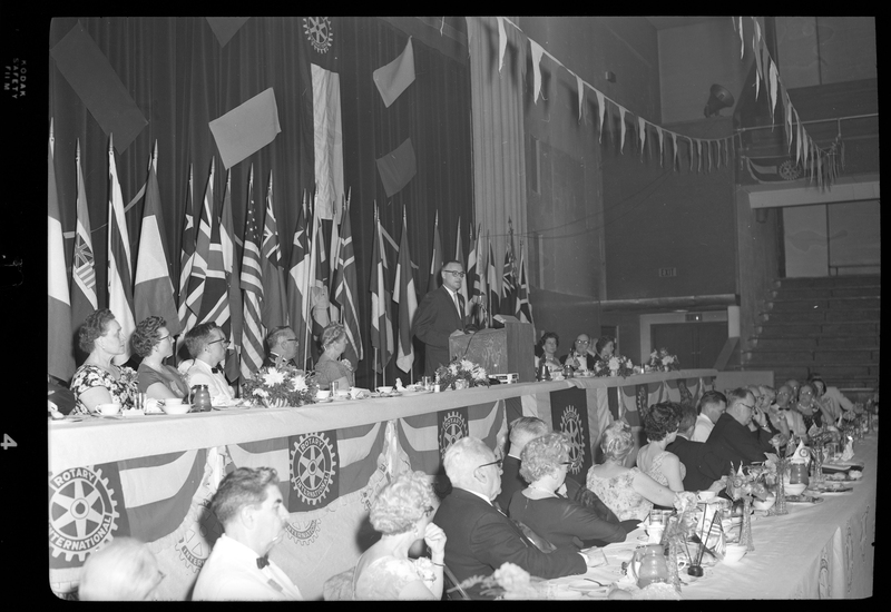 Rotary International Club Convention panel that includes Gordon Beaton, Greek Wells, Guy M'Loughlin, Bill Featherstone, and Bob Green. Various unidentified men and women are seated in two rows with dinner ware on the tables in front of them, looking to an unidentified man who is standing behind a podium. There are banners hanging from the ceiling and various flags along the back wall.