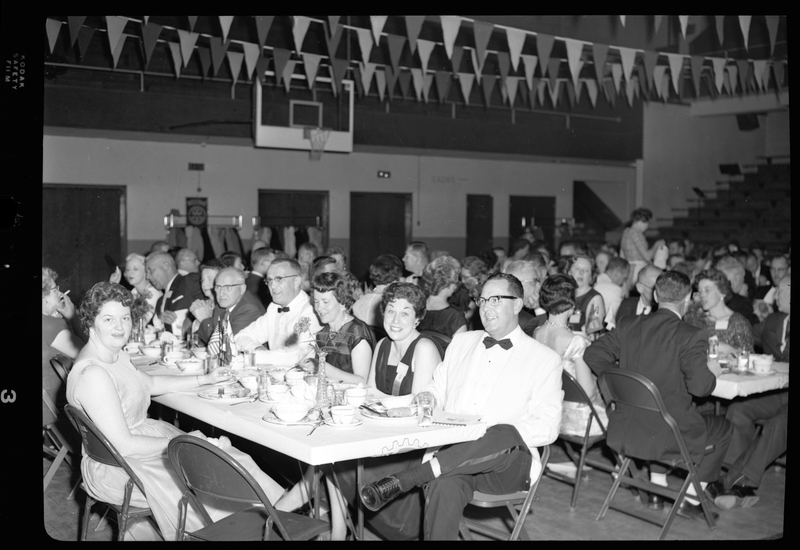 Part of a crowd of unidentified men and women eating at the Rotary International Club Convention. Two women are looking at the camera while the rest of the crowd are interacting with each other. Banners hang across the ceiling and there is a basketball hoop in the background.
