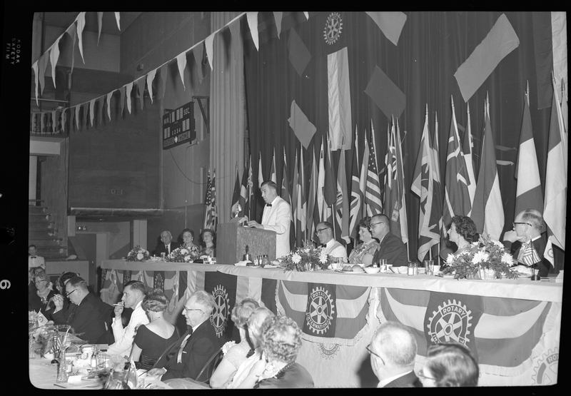 Rotary International Club Convention panel that includes Gordon Beaton, Greek Wells, Guy M'Loughlin, Bill Featherstone, and Bob Green. Various unidentified men and women are seated in two rows with dinner ware on the tables in front of them, looking to an unidentified man who is standing behind a podium. There are banners hanging from the ceiling and various flags along the back wall.