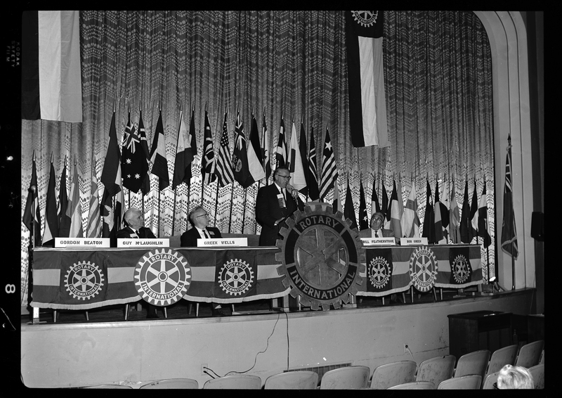 Rotary International Club Convention panel that includes Gordon Beaton, Greek Wells, Guy M'Loughlin, Bill Featherstone, and Bob Green. Bob Green and Gordon Beaton are not behind their name placards, and there is a man addressing the crowd at the podium. There are various flags along the back wall.