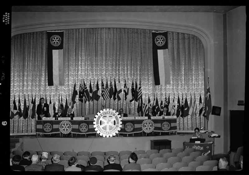 Rotary International Club Convention panel that includes Gordon Beaton, Greek Wells, Guy M'Loughlin, Bill Featherstone, and Bob Green. Some of the men listed are not behind their name placards, and there is an unidentified man speaking to the crowd. There are various flags along the back wall.