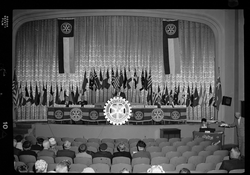 Rotary International Club Convention panel that includes Gordon Beaton, Greek Wells, Guy M'Loughlin, Bill Featherstone, and Bob Green. Some of the men listed are not behind their name placards, and there is an unidentified man speaking to the crowd. There are various flags along the back wall.