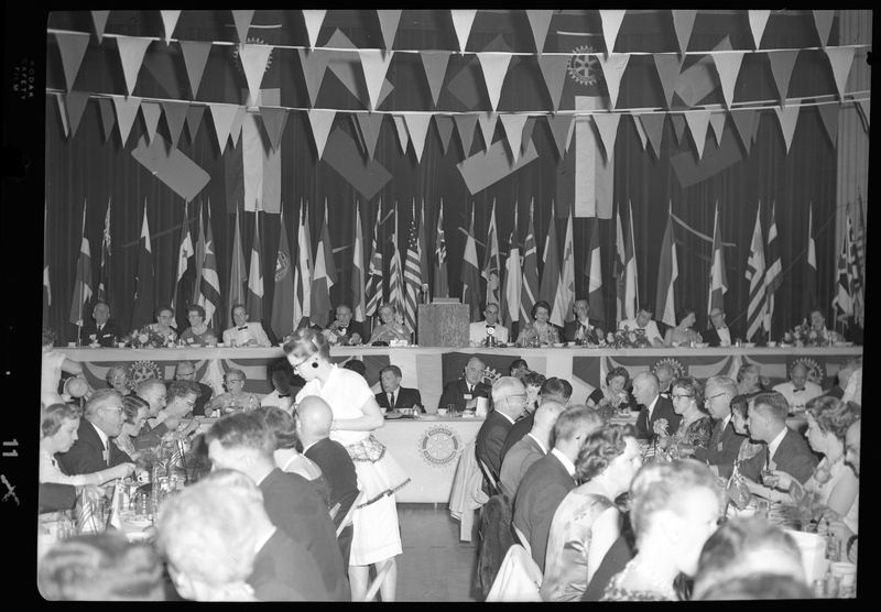 Rotary International Club Convention panel that includes Gordon Beaton, Greek Wells, Guy M'Loughlin, Bill Featherstone, and Bob Green. Various men and women are seated in two rows with dinner ware on the tables in front of them facing a crowd of other dinner guests. There are banners hanging from the ceiling and various flags along the back wall.