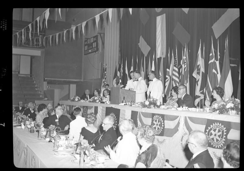 Rotary International Club Convention panel that includes Gordon Beaton, Guy M'Laughlin, Bill Featherstone, Theodore Hopf, Ira Branson, Jack Coventry, and Jack Barre. Various men and women are seated in two rows with dinner ware on the tables in front of them. There are banners hanging from the ceiling and various flags along the back wall. Three men are standing in front of a podium.