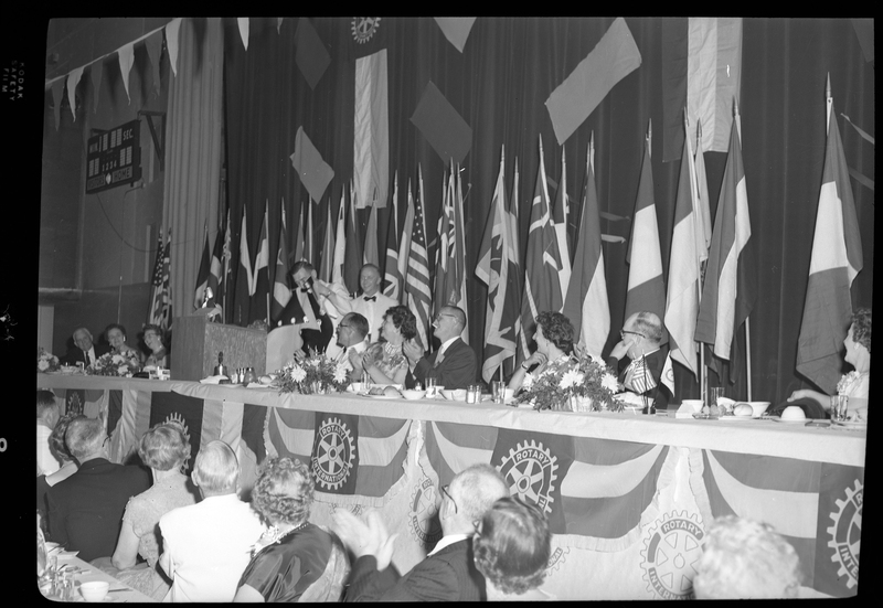 Rotary International Club Convention panel that includes Gordon Beaton, Guy M'Laughlin, Bill Featherstone, Theodore Hopf, Ira Branson, Jack Coventry, and Jack Barre. Various men and women are seated in two rows with dinner ware on the tables in front of them. There are banners hanging from the ceiling and various flags along the back wall. Two men are standing in front of a podium.