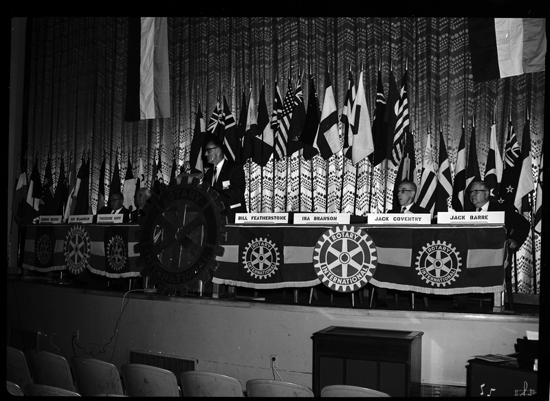 Rotary International Club Convention panel that includes Gordon Beaton, Guy M'Loughlin, Bill Featherstone, Theodore Hopf, Ira Branson, Jack Coventry, and Jack Barre. Some of the people listed are not behind their name placards, and there is an unidentified man speaking to the crowd. There are various flags along the back wall.
