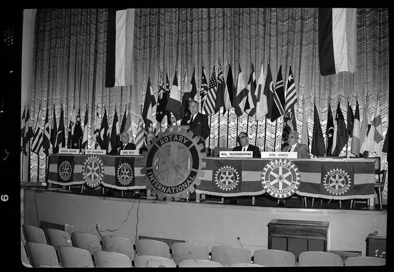 Rotary International Club Convention panel that includes Gordon Beaton, Guy M'Loughlin, Bill Featherstone, Theodore Hopf, Ira Branson, Jack Coventry, and Jack Barre. Some of the people listed are not behind their name placards, and there is an unidentified man speaking to the crowd. There are various flags along the back wall.
