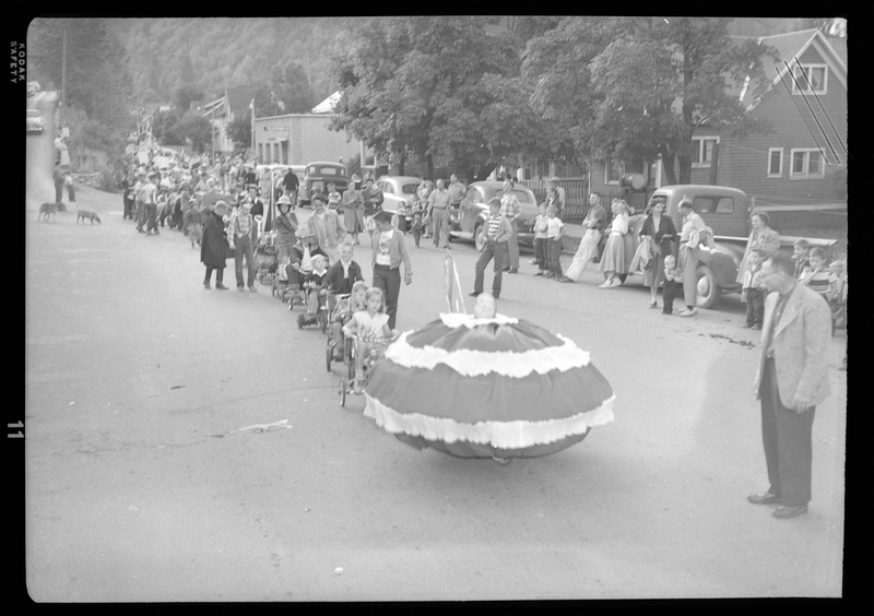 Beginning of the Benevolent and Protective Order of Elks parade. A line of people can be seen down the entire street consisting mostly of children. People can be seen standing on the side of the road watching the parade, with cars, trucks, houses, and dogs in the background.