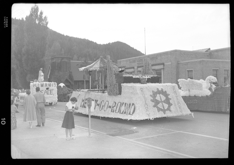 Several parade floats for the Benevolent and Protective Order of Elks are visible. One float has a swan design, another has a merry go round, and there are several unidentified people standing on or near the floats. The floats are in front of some buildings.