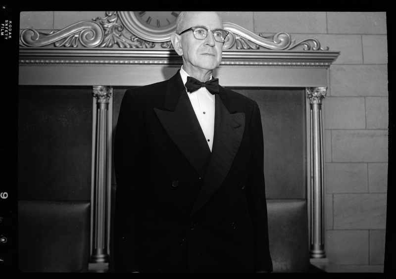 Unidentified man from the Benevolent and Protective Order of Elks Officers standing in a room wearing a tuxedo. There is a clock behind him and he is looking off into the distance. B.P.O.E. Parade (Benevolent and Protective Order of Elks).