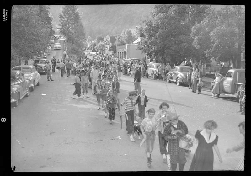 Photo of ongoing Benevolent and Protective Order of Elks Parade where unidentified men, women, and children can be seen progressing down the road while other look on from the sides. Kids can be seen wearing costumes. In the background there are vehicles, trees, buildings, and crowds of people.