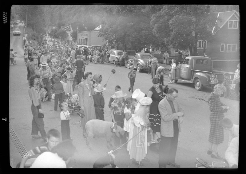 Photo of ongoing Benevolent and Protective Order of Elks Parade where unidentified men, women, and children can be seen progressing down the road while other look on from the sides. Kids can be seen wearing costumes, and there are people holding sheep on leads. In the background there are vehicles, trees, buildings, and crowds of people.