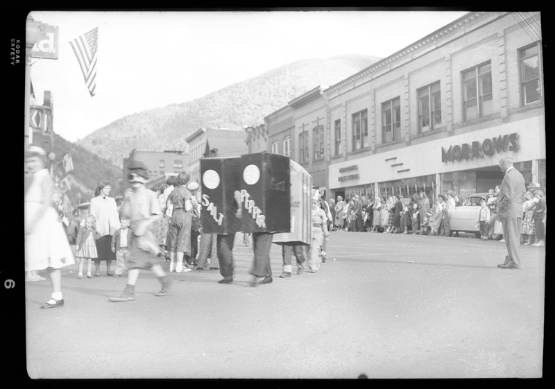 Two people wearing enlarged salt and pepper shaker costumes for the Benevolent and Protective Order of Elks parade. Other people can be seen walking in front of and behind the salt and pepper shakers, as well as a crowd lined up in front of a building to watch the parade.