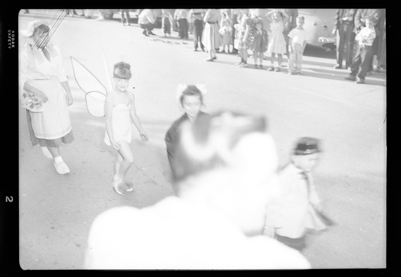 Photo of ongoing Benevolent and Protective Order of Elks Parade where a child in a fairy costume can be seen. There are people standing in the background watching the parade, and the back of a man's head blocks part of the camera.