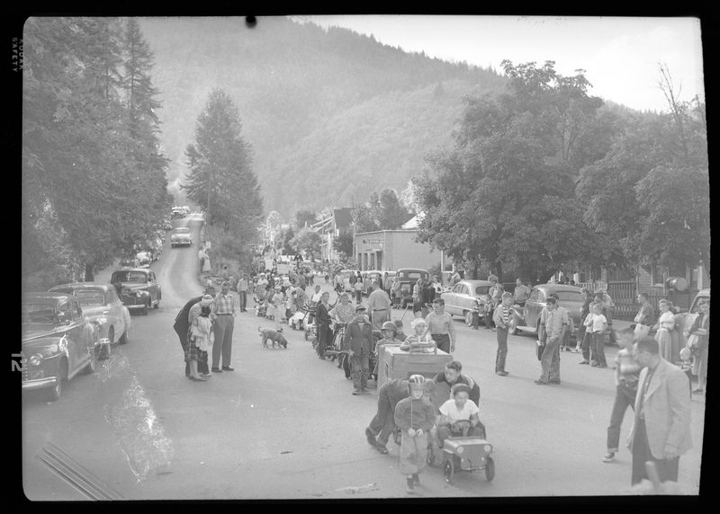 A line of children and adults for the Benevolent and Protective Order of Elks parade. Most children are wearing costumes as they walk or bike down the parade line. In the background people, cars, buildings, dogs, and trees can be seen.