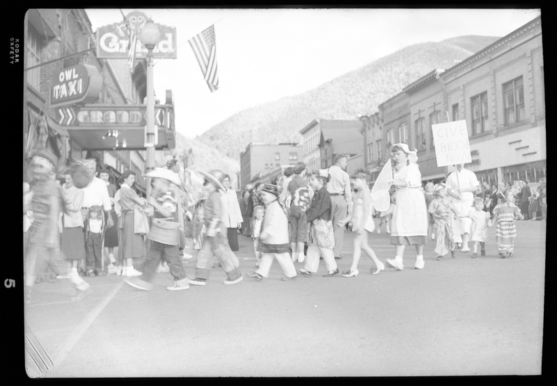 Turning point of the Benevolent and Protective Order of Elk parade where people can be seen turning. In the parade line are children and some adults in costumes, and in the background crowds of people and buildings can be seen.