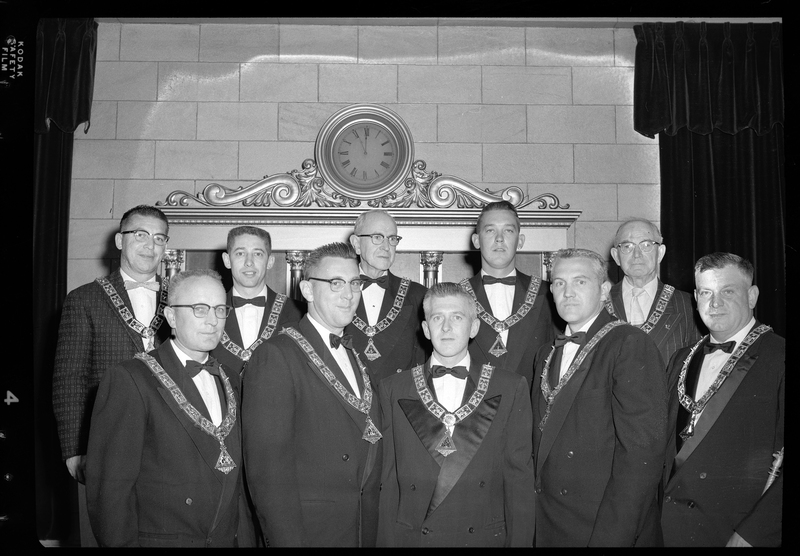 A group of ten men from the Benevolent and Protective Order of Elks Officers wearing matching pendants standing in front of a wall with a clock on it. All of the men are wearing suits or tuxedos. B.P.O.E. Parade (Benevolent and Protective Order of Elks).