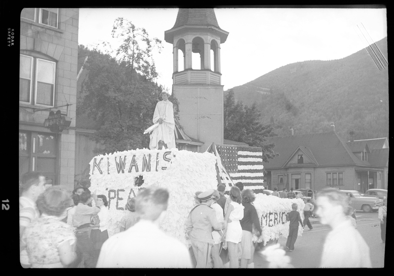 There is a woman standing on top of a parade float along with an American flag for the Benevolent and Protective Order of Elks. There are people crowded around the float and there are buildings, cars, and trees in the background.