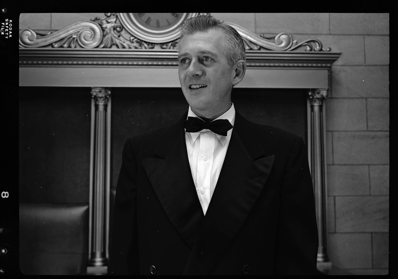 Unidentified man from the Benevolent and Protective Order of Elks Officers standing in a room wearing a tuxedo. There is a clock behind him and he is looking off into the distance.