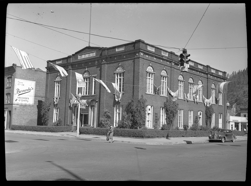 Photo of the building for the Benevolent and Protective Order of Elks taken across a corner street. Flags are hung across the street under the stop light, and there is a car and a woman in front of the building. 