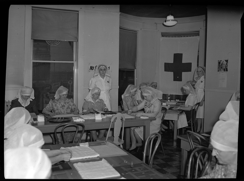 Photo of a group of Red Cross Nurses sitting at tables together. All of the nurses are wearing caps and working on something at the tables. There is a Red Cross Flag hanging over one of the windows in the room.