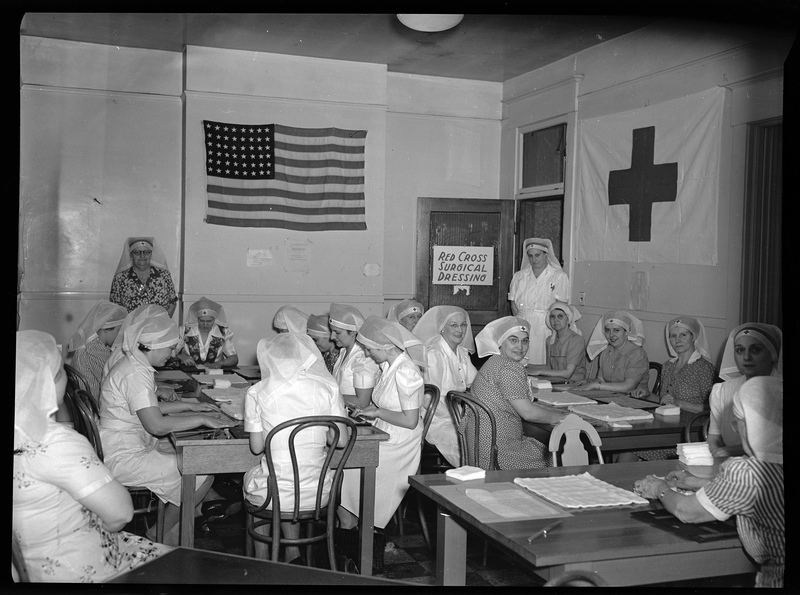 Photo of a group of Red Cross Nurses sitting at tables together, some working and some looking at the camera. There is a Red Cross Flag and an American flag hanging on the walls. Two women are standing, one of which is by the door with a sign that reads "Red Cross Surgical Dressing."
