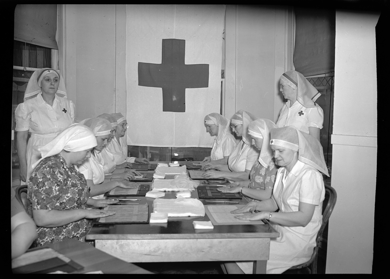 Photo of a group of Red Cross Nurses sitting at table together, with two women standing on either side of the table looking over the others. All of the nurses are wearing caps and working on something at the table. There is a Red Cross Flag hanging over one of the windows.
