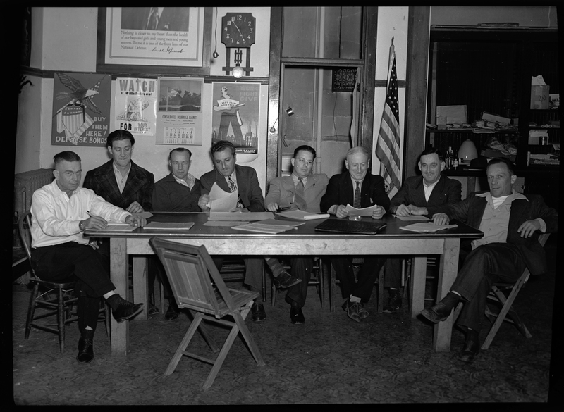 A group of men sitting around a table together, looking at various papers. There are posters, a clock, and an American flag on the wall behind them, as well as an empty chair in front of the table. Some of the men are looking at the camera.