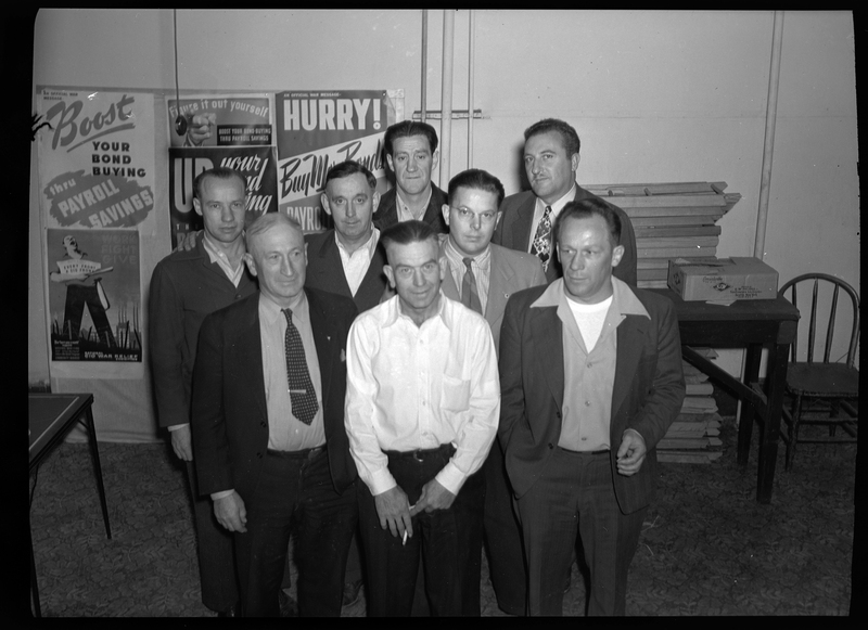 A group of men standing together while posing for a photo. There are posters on the wall behind them, as well as a table and chair. Some of the men are looking at the camera while others are looking elsewhere.