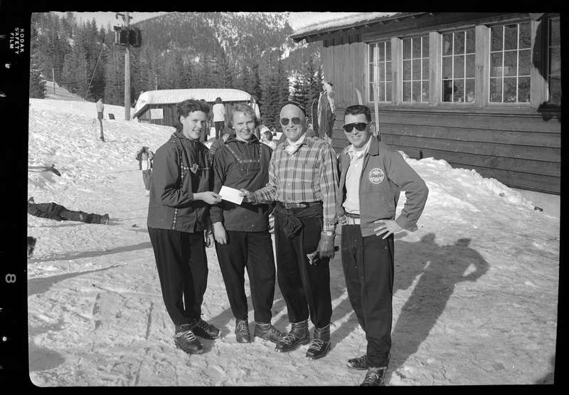 Photo of four unidentified people posing for a photo at ski school. There are a few buildings, people, and trees in the background. There is snow covering the ground, trees, and roofs.