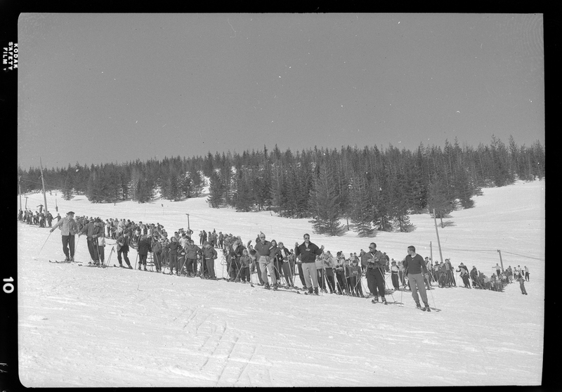 Photo of a large group of people, mostly children, standing in the snow at ski school. Everyone is wearing skis, and there are trees in the background.