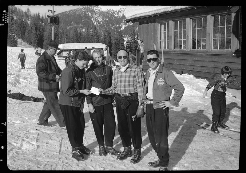 Photo of four unidentified people posing for a photo at ski school, with a man walking behind them and a child moving by on skis. There are a few buildings, people, and trees in the background. There is snow covering the ground, trees, and roofs.