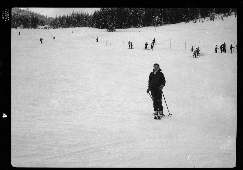 Photo of a woman moving down the snow covered hill on skis. In the background you can see some adults and children either skiing down the hill or being pulled up by the lift. There are snow covered trees visible in the background.