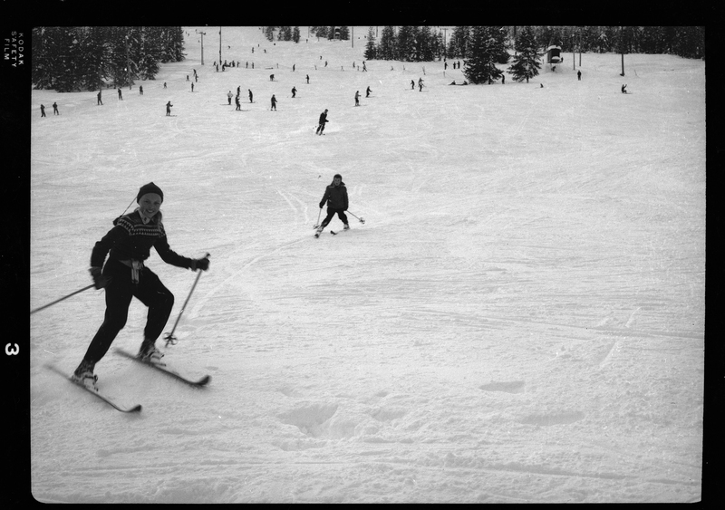 Photo of two children moving down a snow covered hill on skis, one child much closer to the camera than the other. In the background there are other people skiing down the hill, as well as snow covered trees and a ski lift.