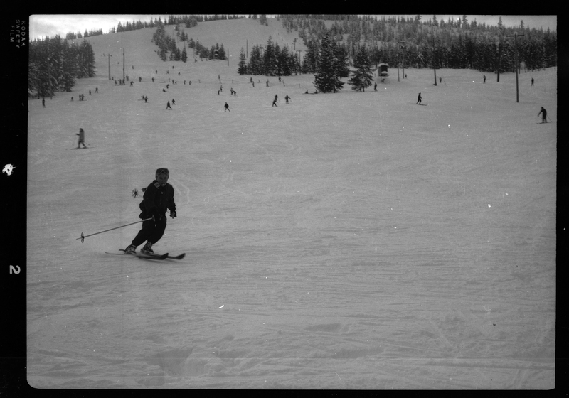 Photo of a child moving down a snow covered hill on skis. In the background there are other people skiing down the hill, as well as snow covered trees and a ski lift.