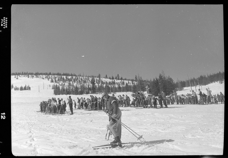 Photo of a man wearing skis, with a large group of people lined up in several rows in the background. There are trees and a ski lift visible behind the man and the crowd.