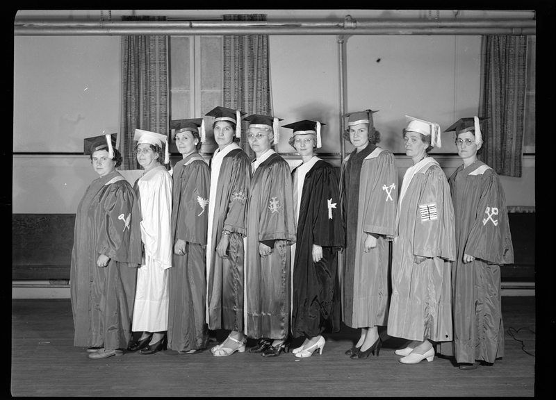 Nine women standing in a line, heads turned to the camera, and all wearing graduation caps and gowns. They are standing in a room.