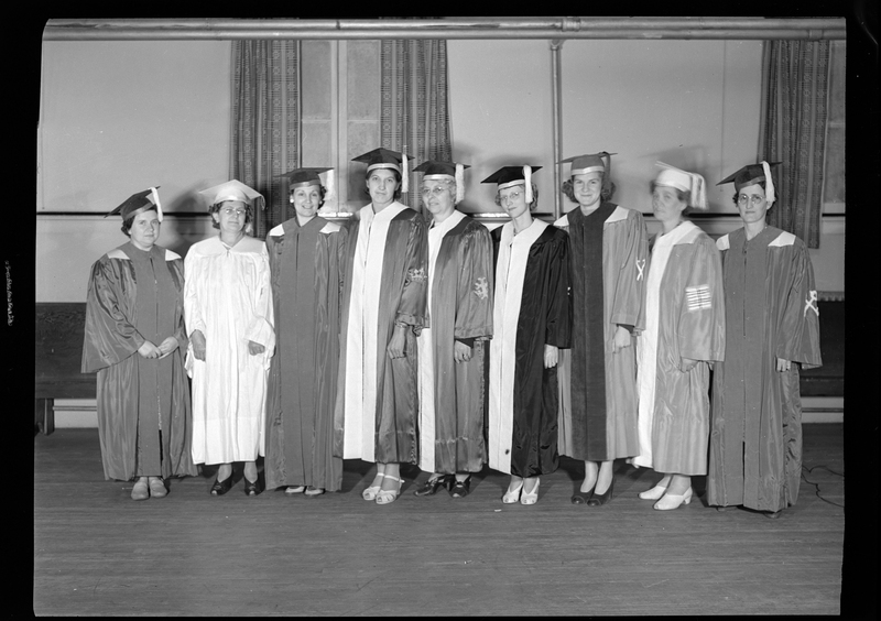 Nine women standing in a line, facing the camera, and all wearing graduation caps and gowns. They are standing in a room.