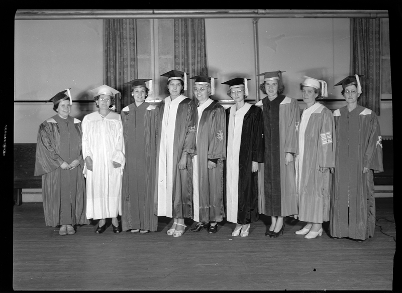 Nine women standing in a line, facing the camera with some smiling, and all wearing graduation caps and gowns. They are standing in a room.